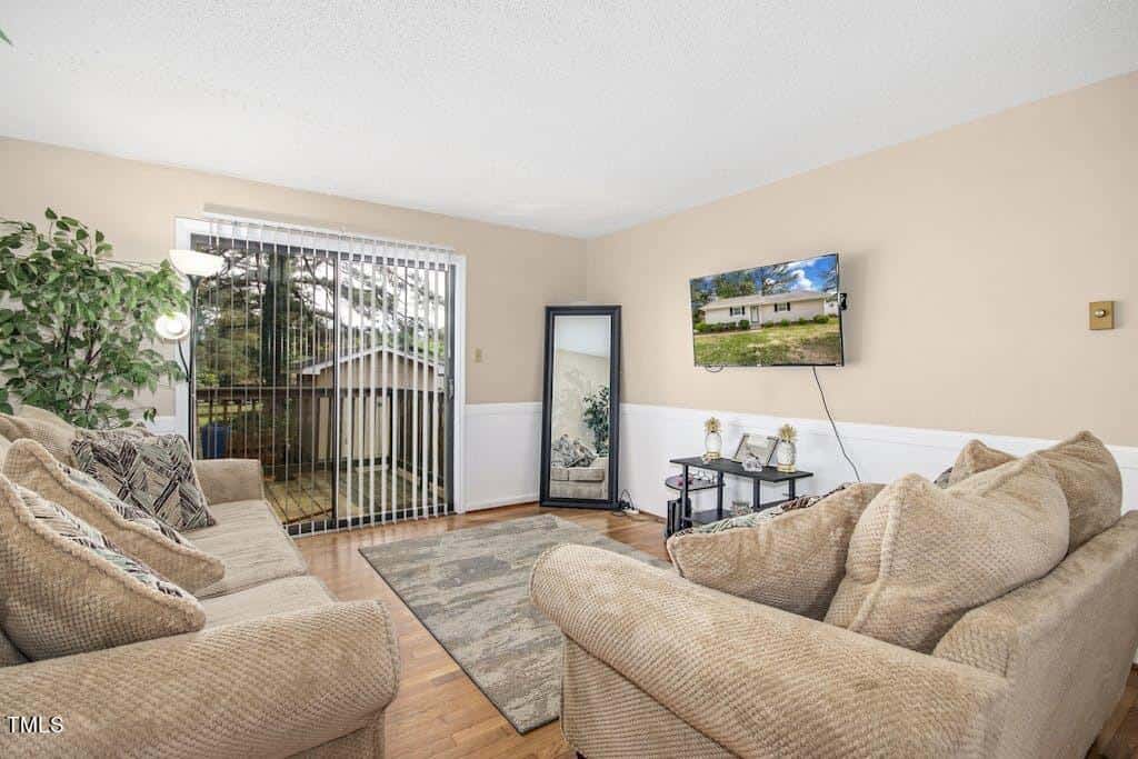 A cozy living room featuring two beige sofas, a patterned rug, and a coffee table. there is a sliding glass door leading to an outdoor area and a large framed landscape picture on the wall.