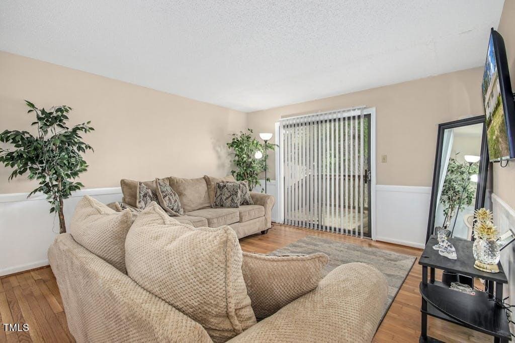 A cozy living room featuring a large beige sectional sofa, hardwood floors, and a sliding glass door with vertical blinds. decor includes a potted plant and wall-mounted flat-screen tv.