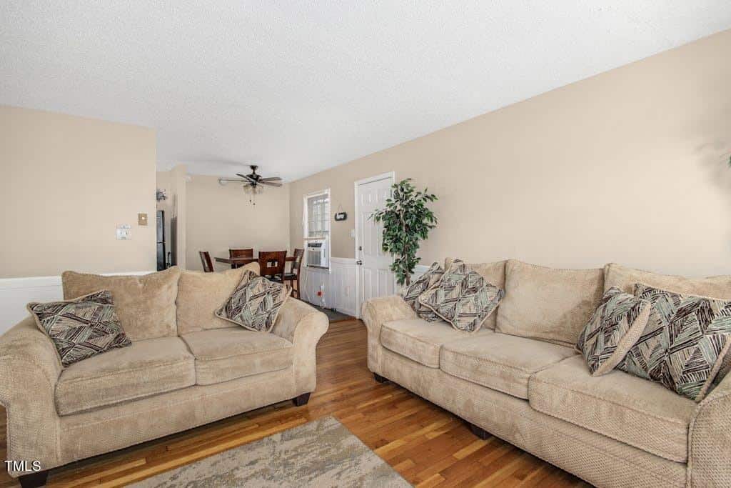 A cozy living room with two beige sofas adorned with patterned cushions, a small rug, hardwood floors, and a ceiling fan. the room has a neutral color palette, doors, and windows for natural light.