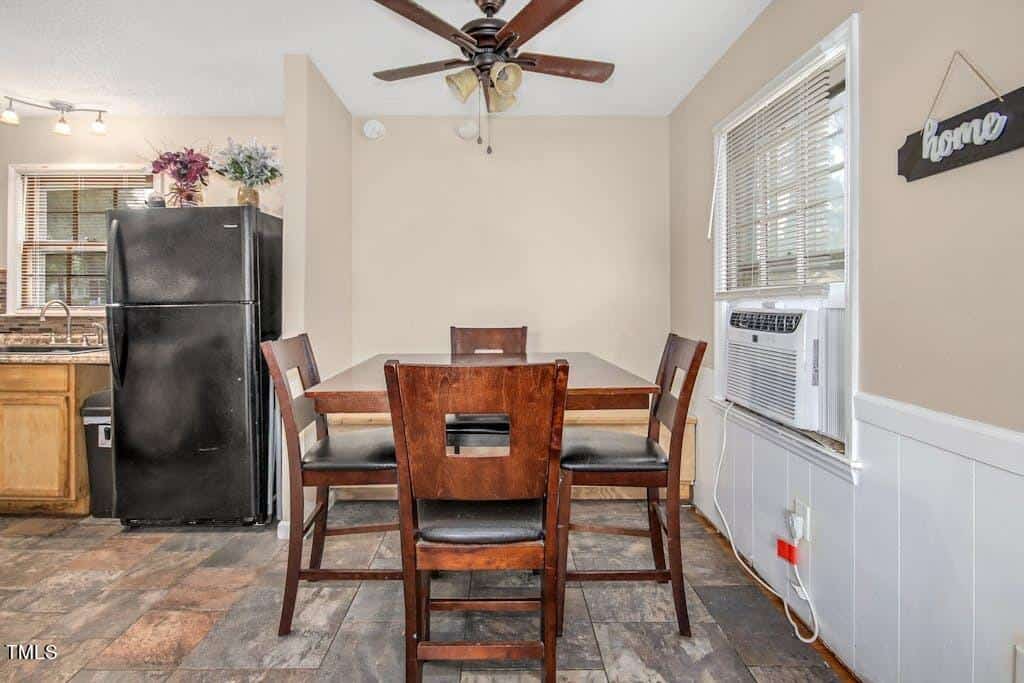 A cozy kitchen with a black refrigerator, a wooden dining table with chairs, cream walls, and a ceiling fan. there is a "home" sign hanging on the wall and a window air conditioner.