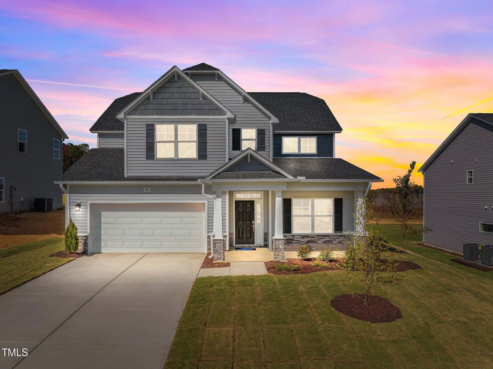 A two-story modern suburban house with gray siding, a double garage, and a covered front porch, sits on a neatly landscaped lawn at sunset with a vibrant purple and yellow sky in the background.