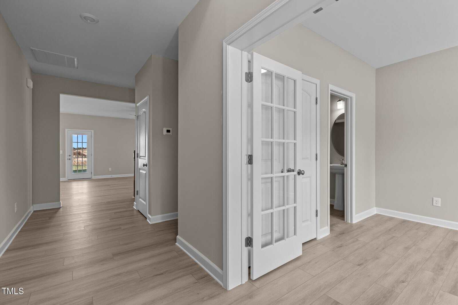 A bright, modern hallway with light wood floors, beige walls, and white trim. French doors open to a small room on the right, and an open doorway leads to a larger, sunlit area in the background.