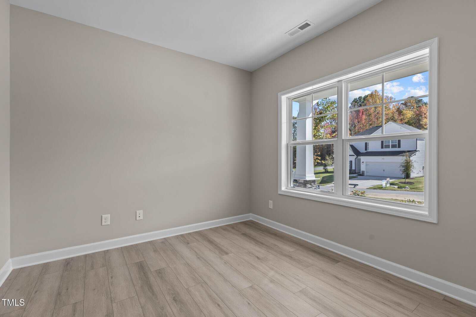 A small empty room with light wood flooring, beige walls, and a large window showing a suburban street with houses, green lawns, and trees. The room has white trim and electrical outlets on one wall.