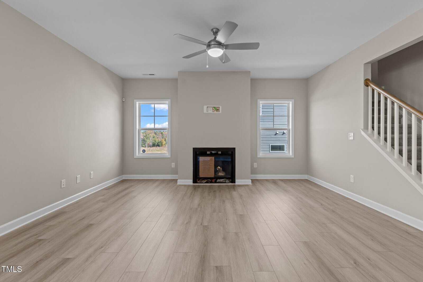 Bright, empty living room with light wood floors, beige walls, a ceiling fan, fireplace centered between two windows, and a staircase with a white railing to the right.