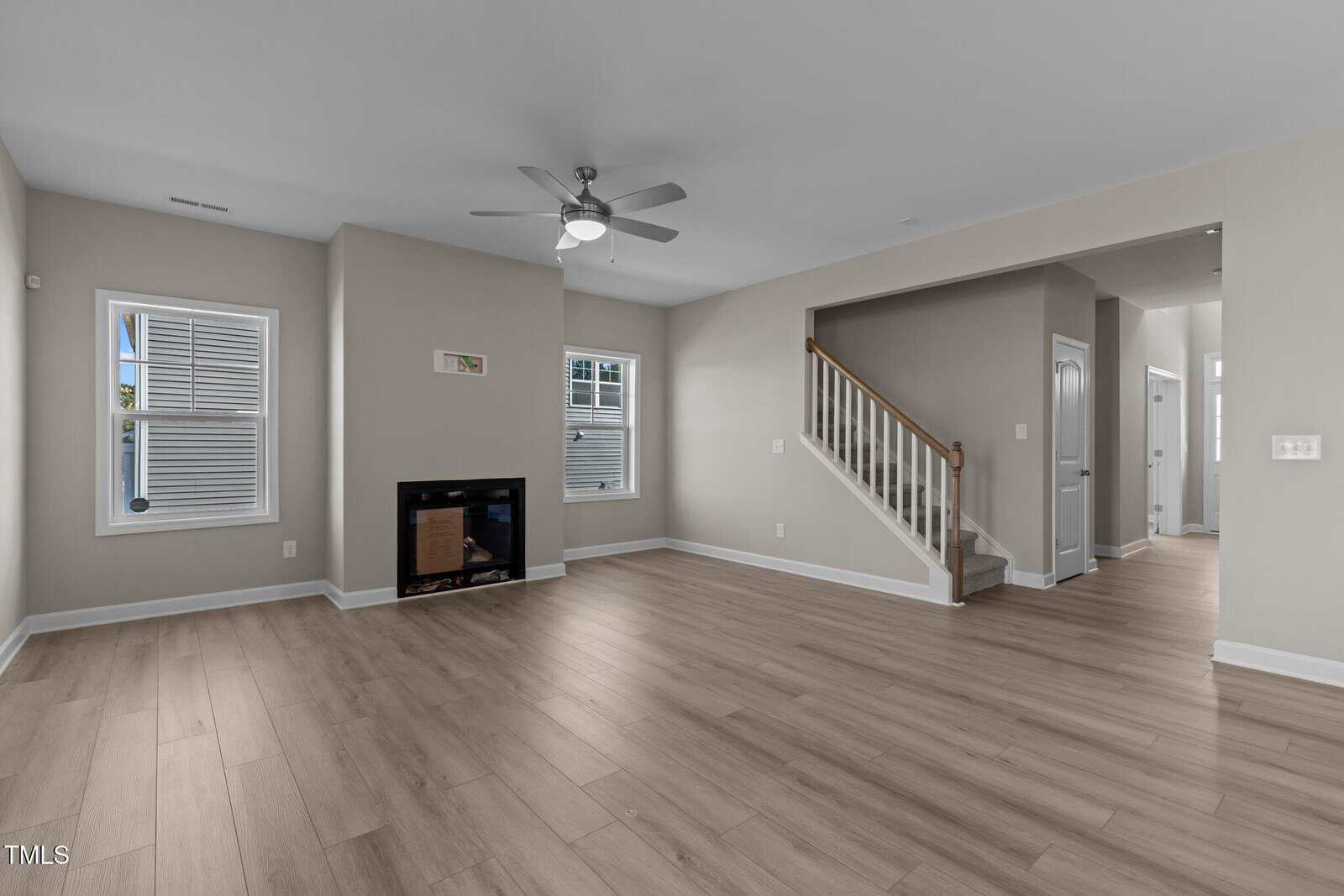 A spacious, empty living room with light wood flooring, beige walls, a ceiling fan, a fireplace, two windows, and a staircase leading upstairs. A hallway is visible to the right of the stairs.