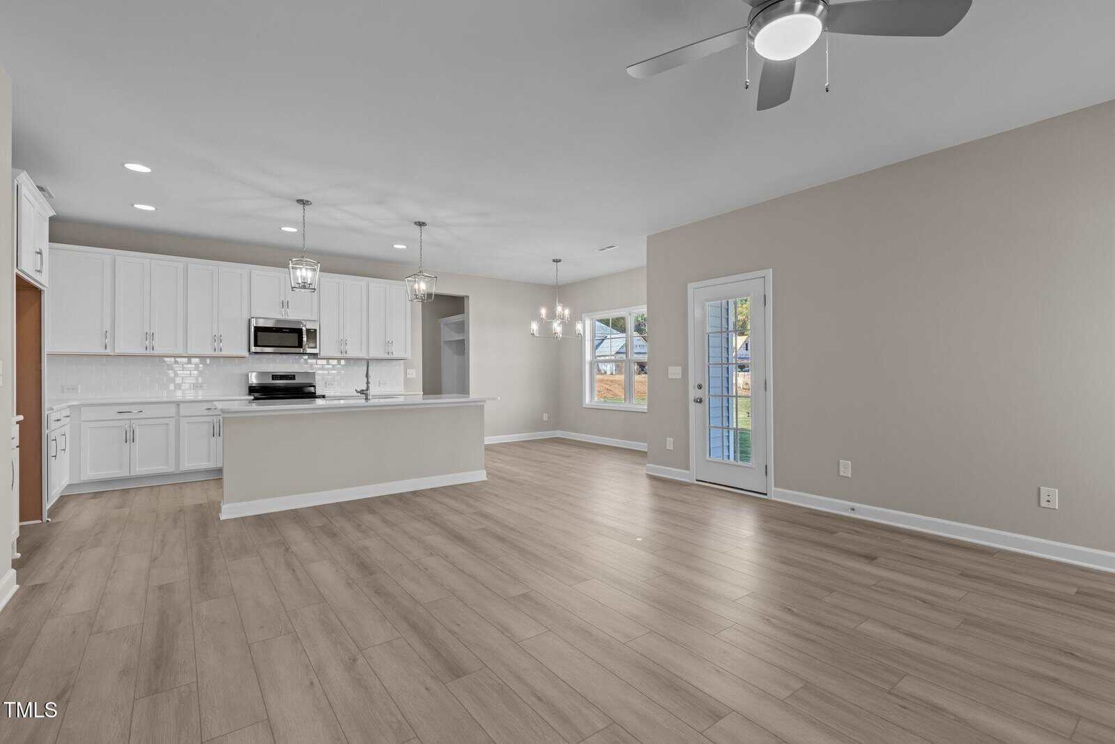 Modern open-concept kitchen and living area with light wood flooring, white cabinets, stainless steel appliances, pendant lighting, and large windows providing natural light. A ceiling fan and a glass door lead outside.