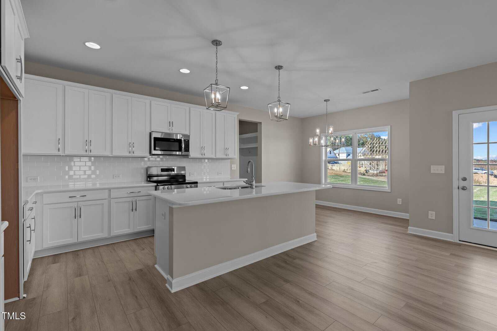 Modern kitchen with white cabinets, stainless steel appliances, a large island with sink, light wood flooring, and a spacious dining area by windows showing a backyard and blue sky through the glass door.