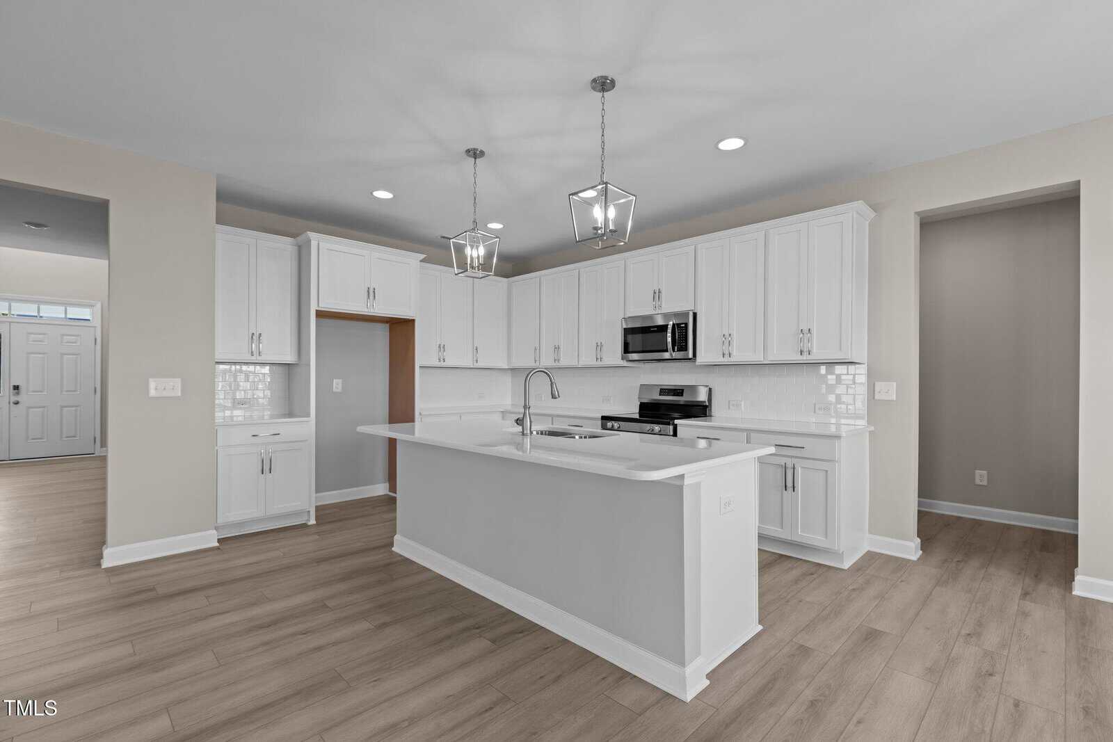 Modern kitchen with white cabinets, stainless steel appliances, a large center island with a sink, pendant lights above, subway tile backsplash, and light wood flooring. Walls are light beige, creating a bright, open space.