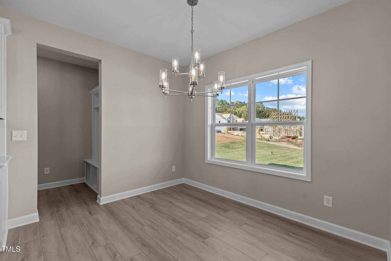 A bright, empty dining room with light wood flooring, beige walls, a modern chandelier, and a large window showing a view of a grassy yard and a house under construction outside.