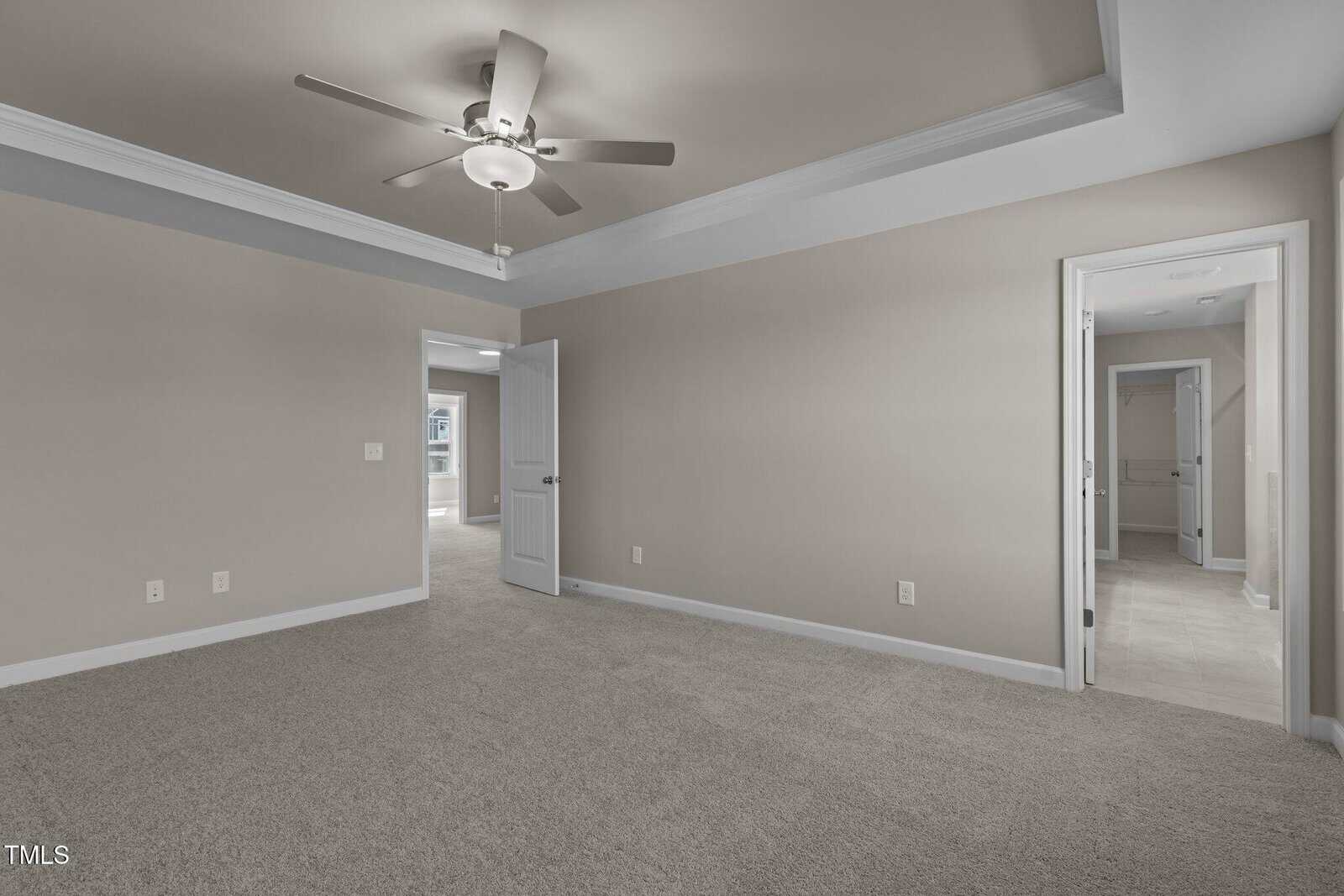 A spacious, empty bedroom with beige walls, carpeted floor, ceiling fan, tray ceiling, an open door, and two doorways leading to tiled bathrooms.