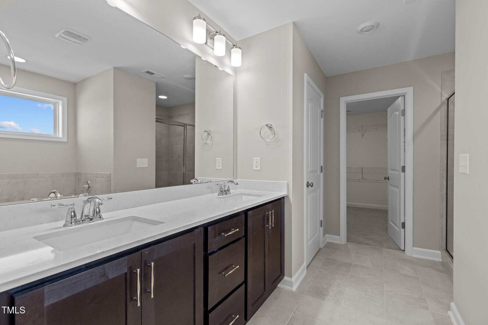 Modern bathroom with a double sink vanity, dark wood cabinets, large wall mirror, neutral tile flooring, and a walk-in closet visible through an open door. Bright lighting and a small window provide natural light.
