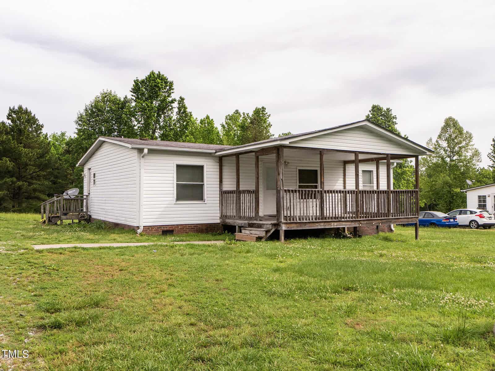 A white single-story house with a wooden porch and ramp sits on a grassy lawn at 575 George Sherman Road in Rougemont. Surrounded by trees, the house is accompanied by parked cars on the right, under an overcast sky.