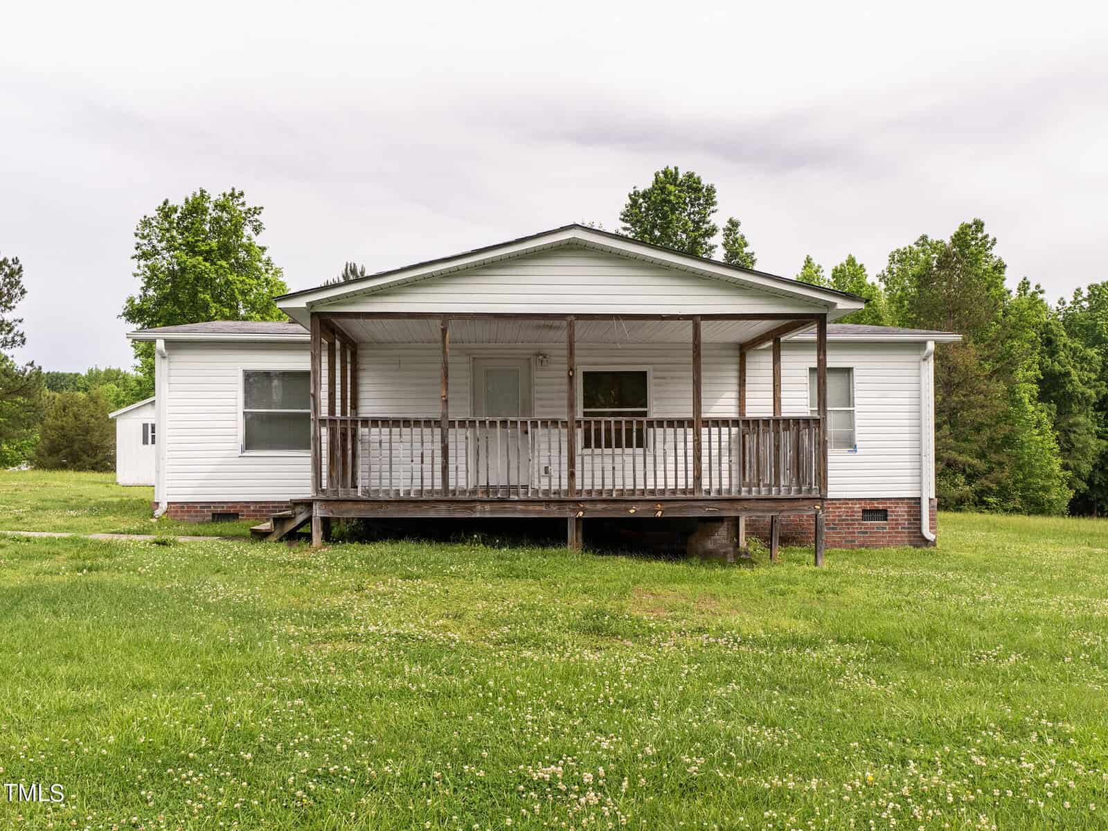 A small white house with a wooden front porch stands on a grassy lawn. The porch has a railing and steps leading down to the yard. The house is surrounded by green trees under a cloudy sky.