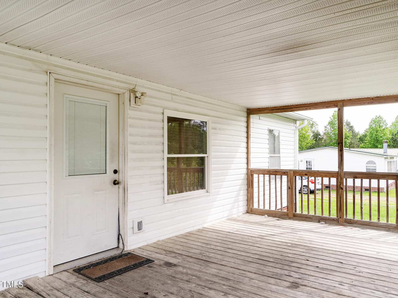 A covered wooden porch with a white door and a window. The porch has wooden railings, a brown floor, and a white ceiling. In the background, there are trees and a parked car on a driveway.