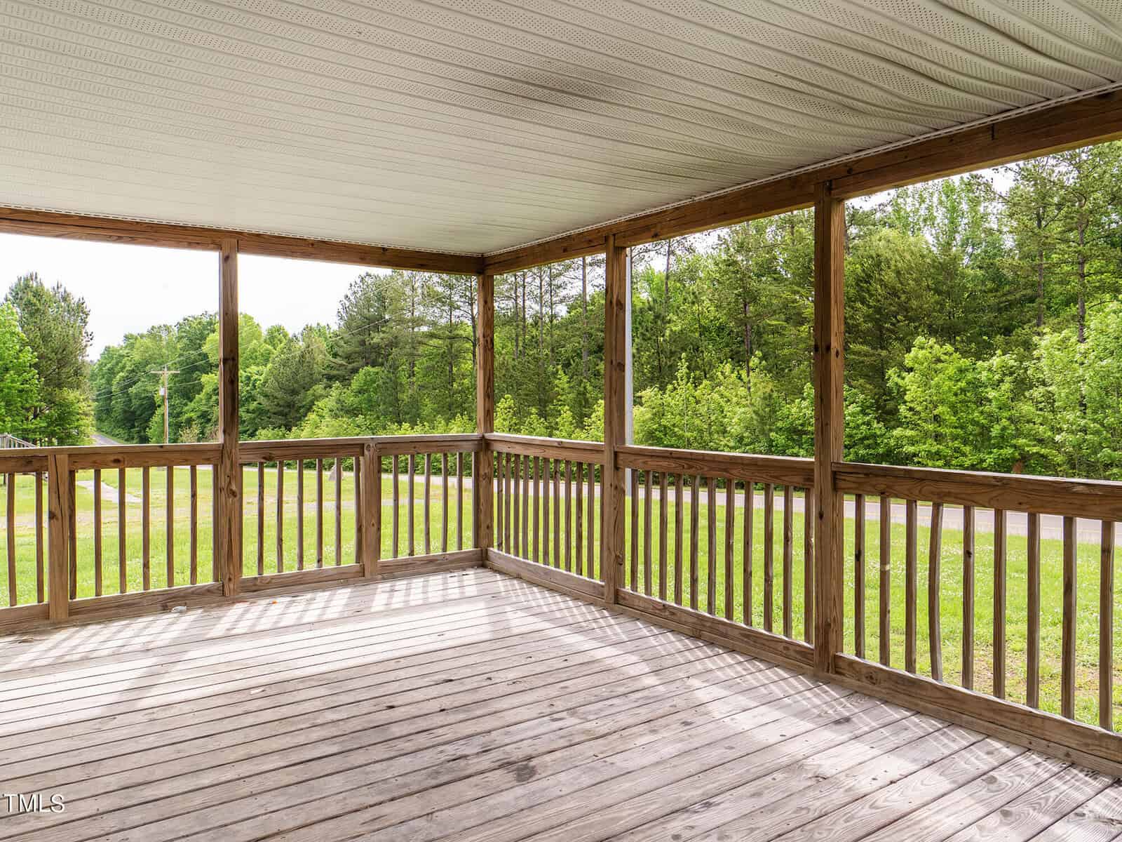 A spacious wooden porch with a railing overlooks a lush green lawn and trees. The porch is covered with a roof, offering a peaceful outdoor space surrounded by nature.