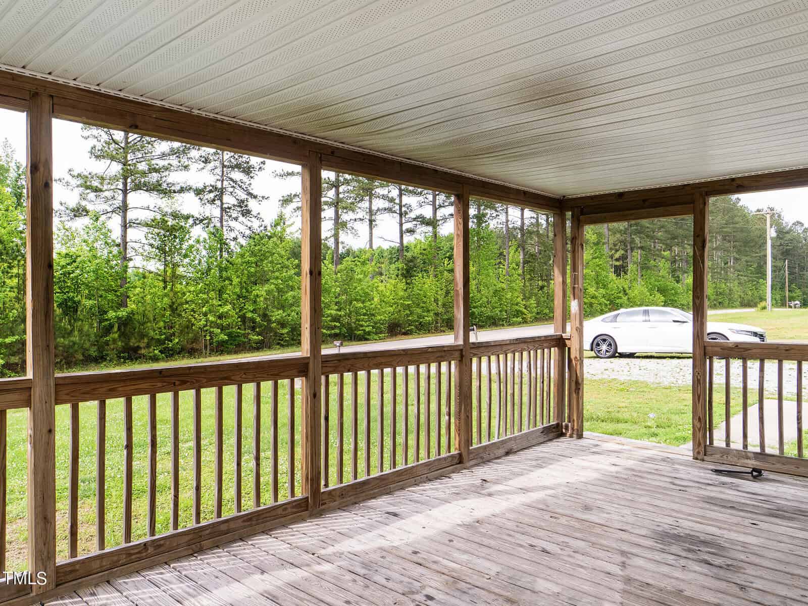 Wooden porch with railing and roof, overlooking a green lawn and trees. A white car is parked on a gravel driveway in the background. The porch provides a scenic view of the surrounding nature.