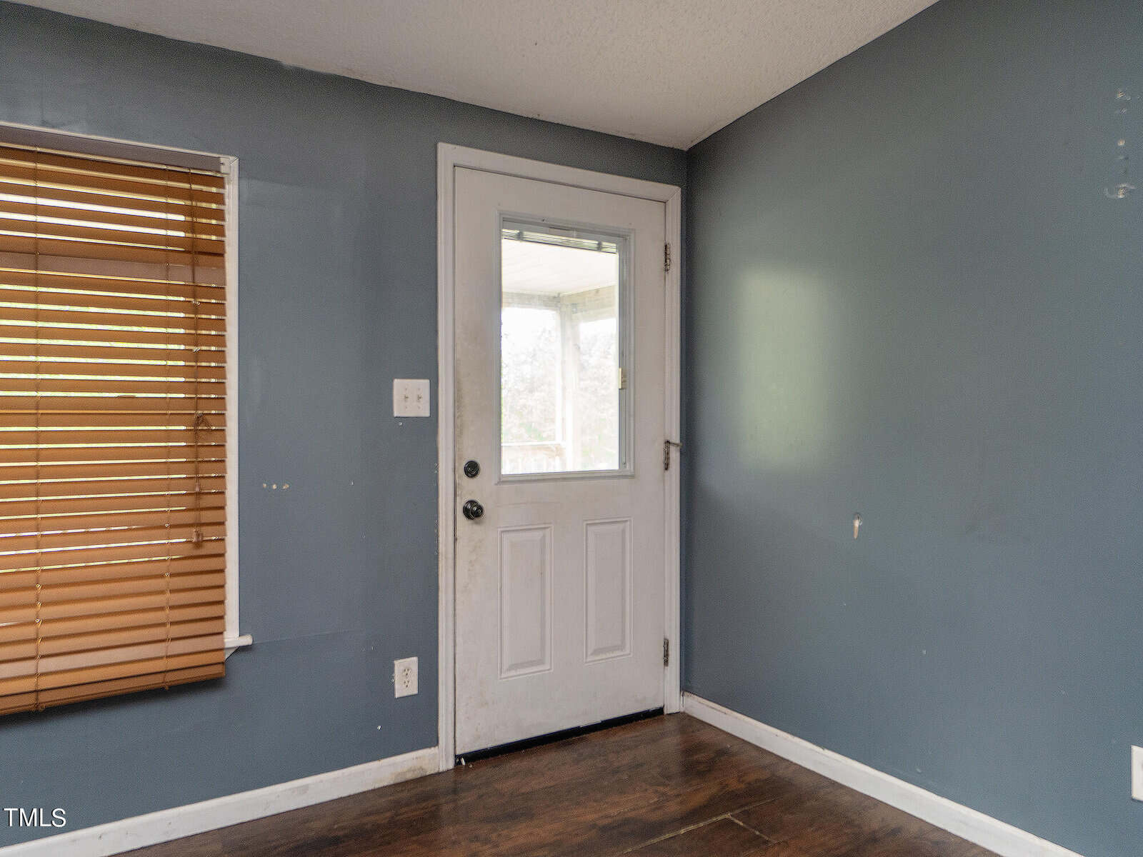 A small room with blue-gray walls and a wooden floor, featuring a closed white door and a window with wooden blinds. The door has a metallic handle and a small window at the top. Light enters softly, brightening the space.