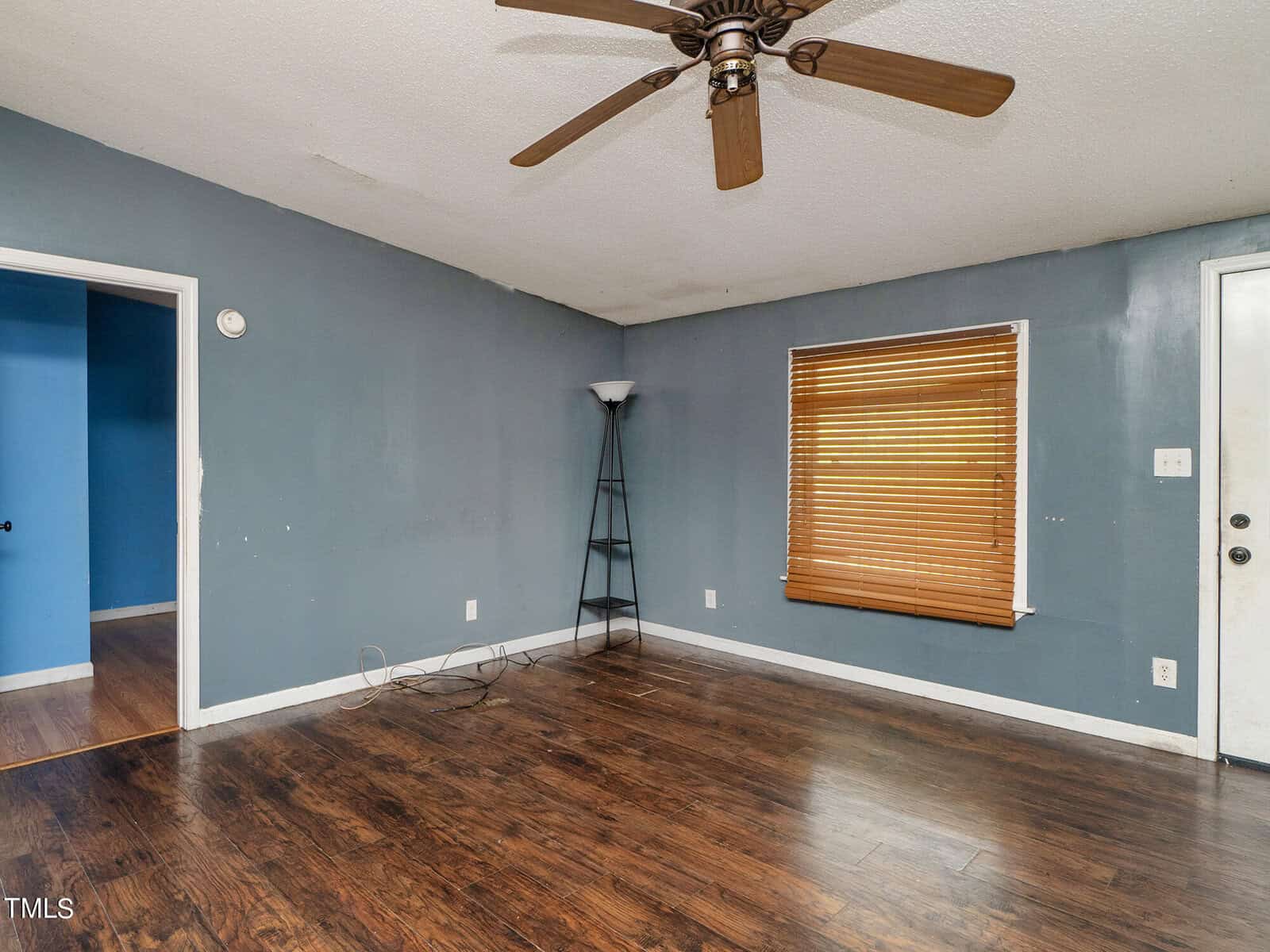 An empty room with dark wooden floors and blue walls. A window with closed blinds is on the right wall. A ceiling fan hangs from the white ceiling. A black floor lamp stands in the corner near a doorway leading to another room.