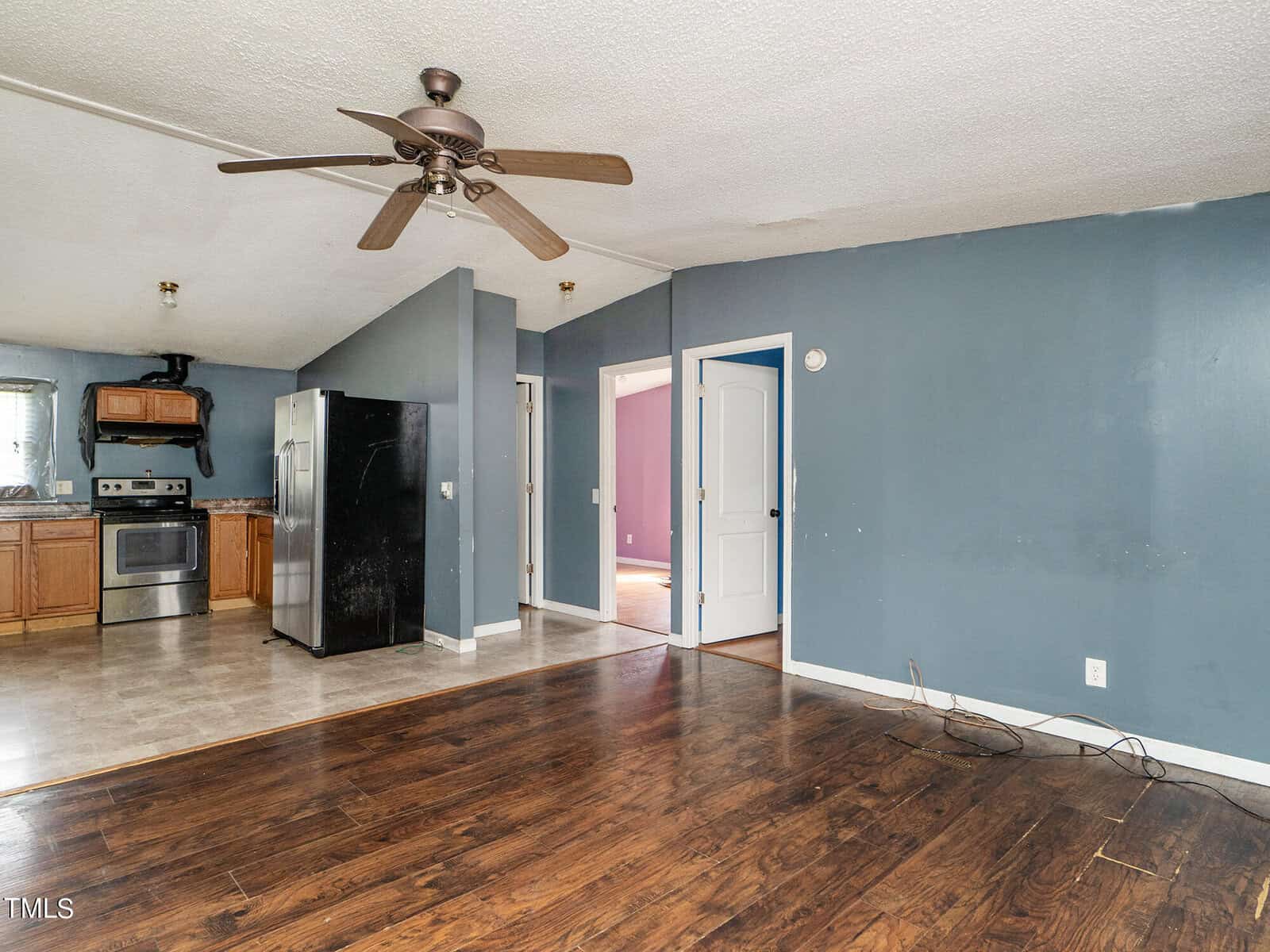 A room with blue walls features dark wood flooring and beige tile. There's a large ceiling fan, a stainless steel oven, a black refrigerator, and wooden cabinets. Two doorways lead to adjacent rooms.
