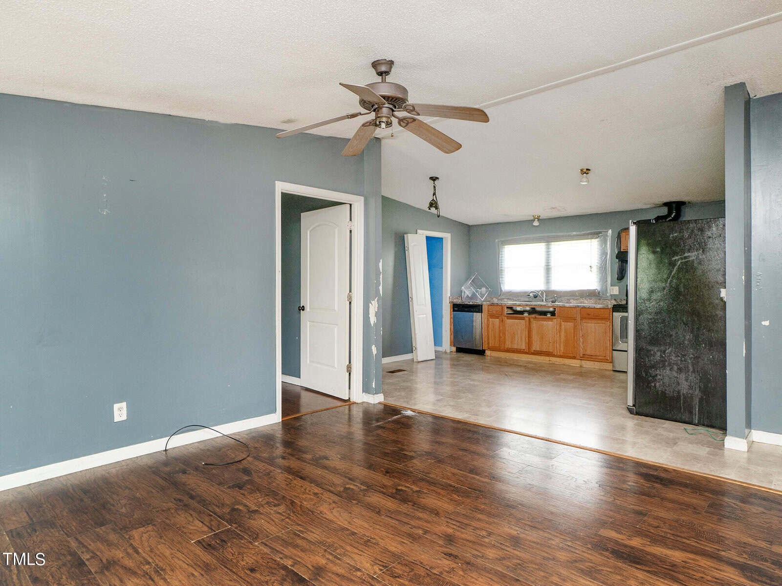 A sparsely furnished room with wooden floors and blue walls, featuring a ceiling fan. The space opens into a kitchen area with tiled flooring, wooden cabinets, and appliances including a fridge and stove. A white door is visible on the left.