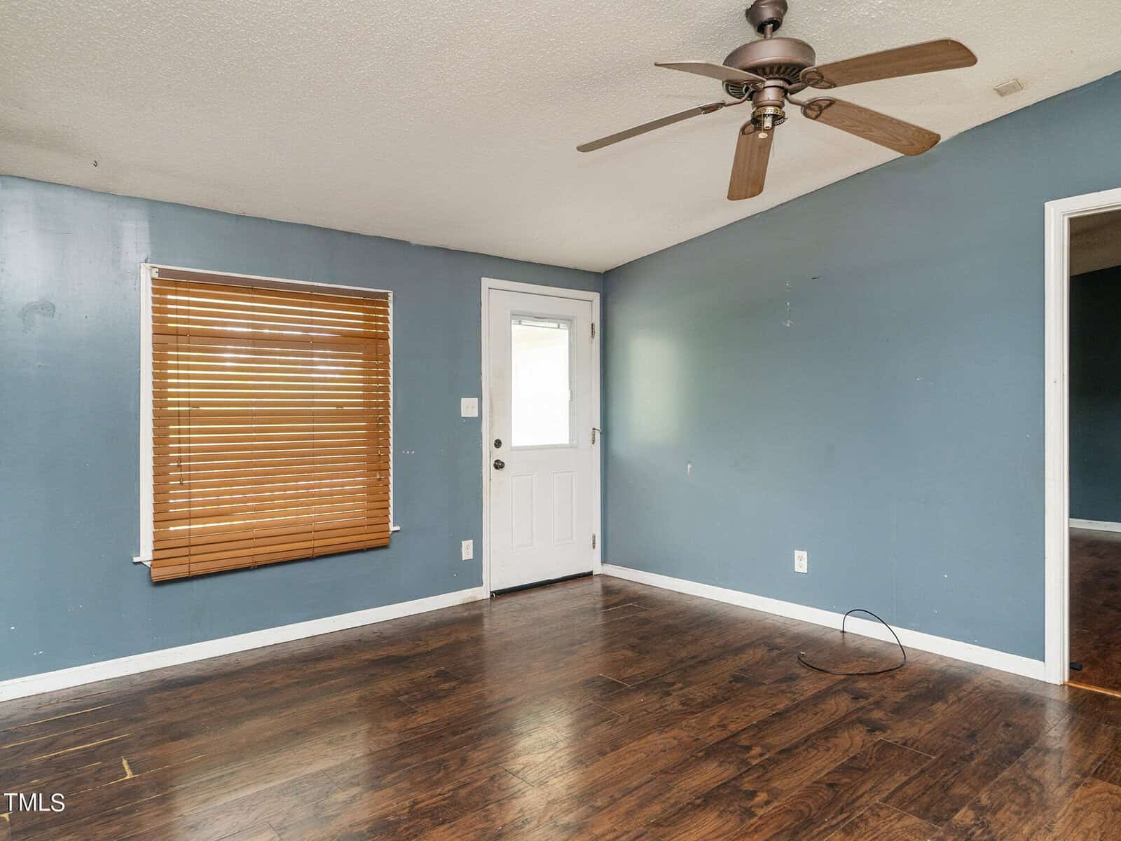 An empty room with blue walls and dark wooden floors. A ceiling fan with wooden blades is mounted on the ceiling. A window with closed blinds is on one wall, and a white door with a frosted glass panel is on another wall.