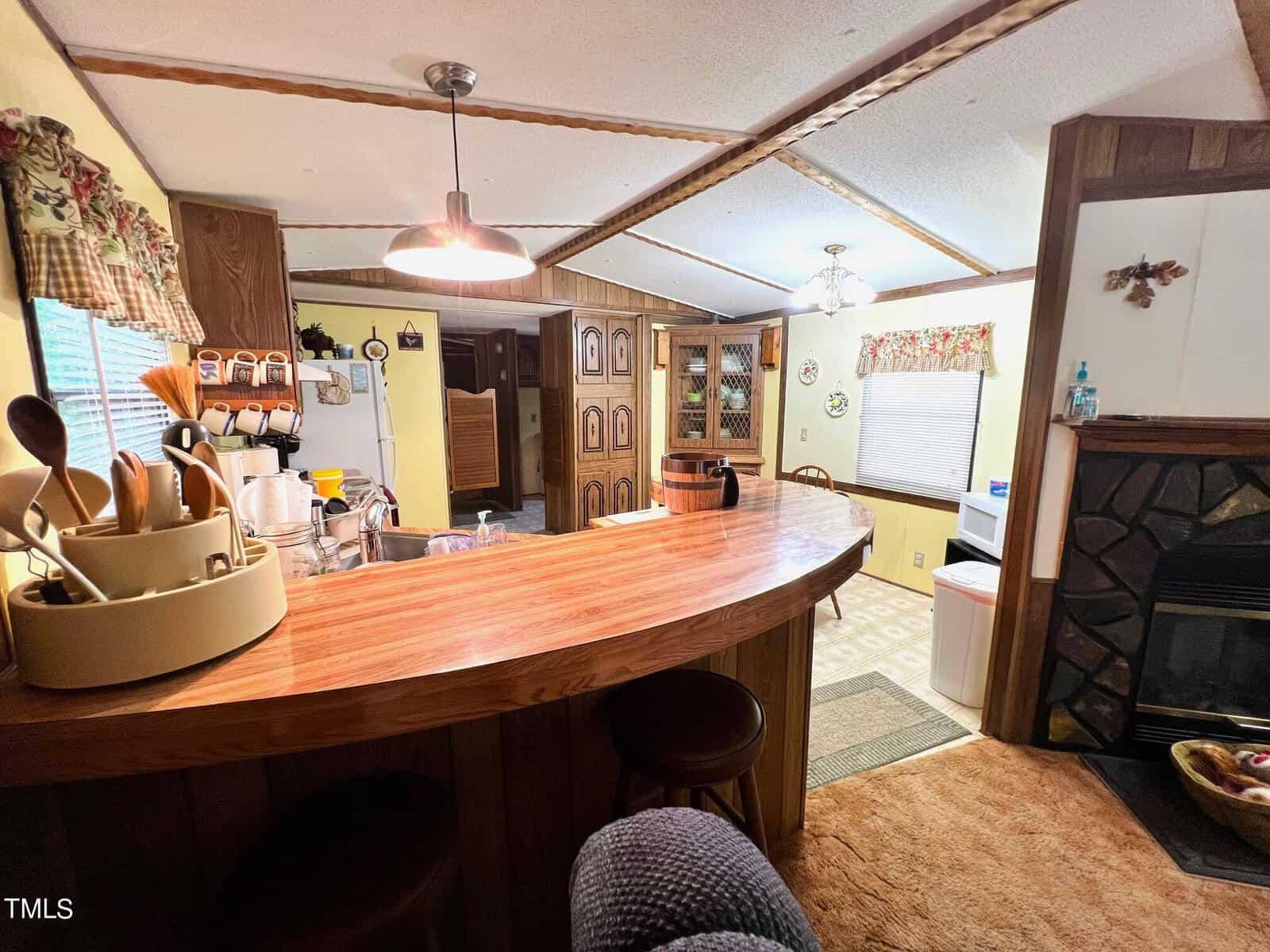 A cozy kitchen with a curved wooden countertop, bar stools, hanging pendant light, and wood-paneled cabinetry. Various kitchen utensils are on the counter. Adjacent is a dining area with a window and decorative curtains.