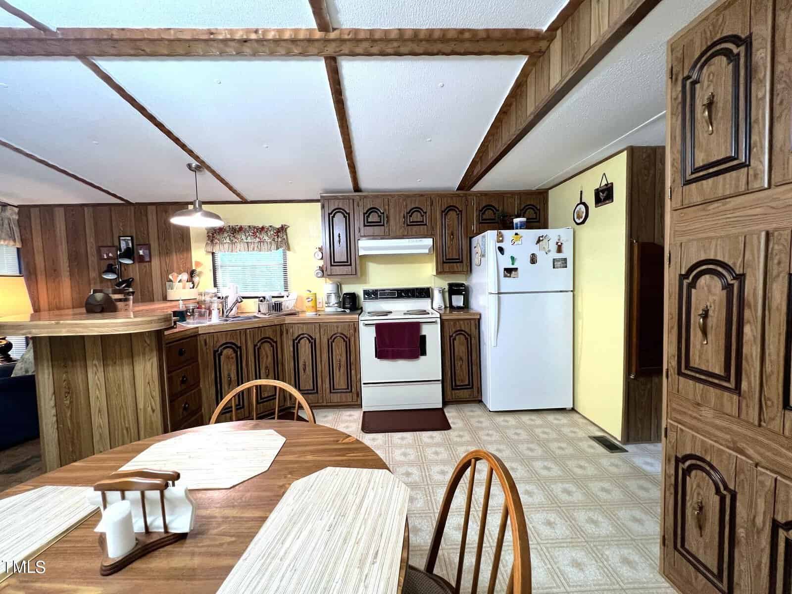 A cozy kitchen with wood-paneled walls and ceiling beams. There's a round dining table with place mats and a condiment set. The kitchen features a stove, refrigerator, and ample cabinets. A small counter with stools divides the kitchen from the main area.
