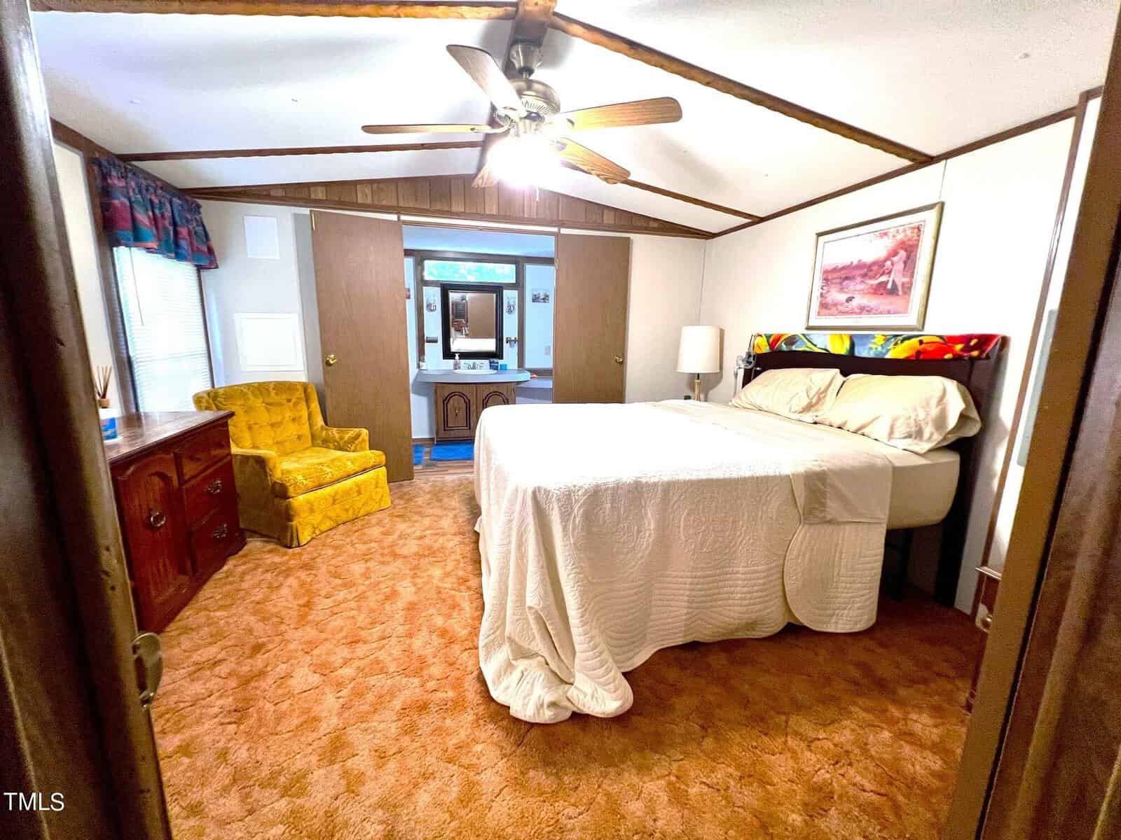 A cozy bedroom with a double bed covered in a white quilt, a wooden dresser with a yellow armchair, a ceiling fan, and a wooden-paneled accent wall with a painting. A doorway leads to an adjoining room with blue carpet.