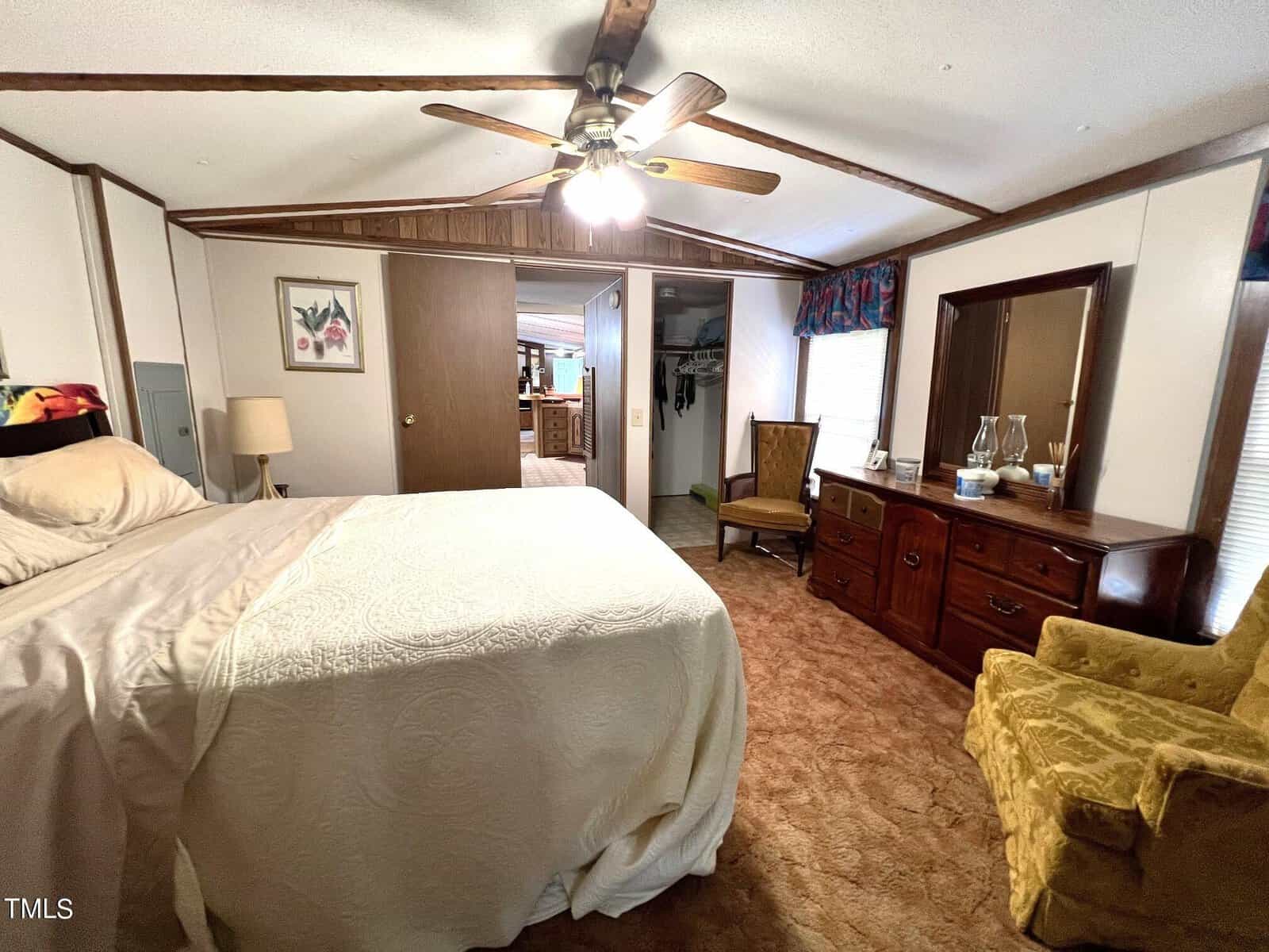 A cozy bedroom features a large bed with white bedding, a ceiling fan, a wooden dresser with a mirror, and a yellow armchair. The room has brown carpet and wood accents, with a view into an adjacent room through an open doorway.