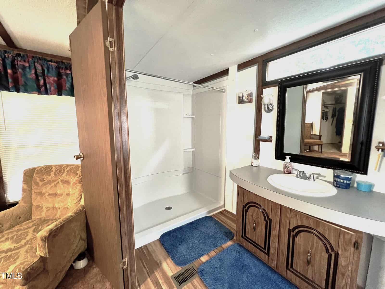 A bathroom with a wooden vanity and large mirror, featuring a white sink with soap dispensers. Adjacent is a white shower with sliding doors. The room has wood accents, blue bath mats, and a beige armchair by the window.