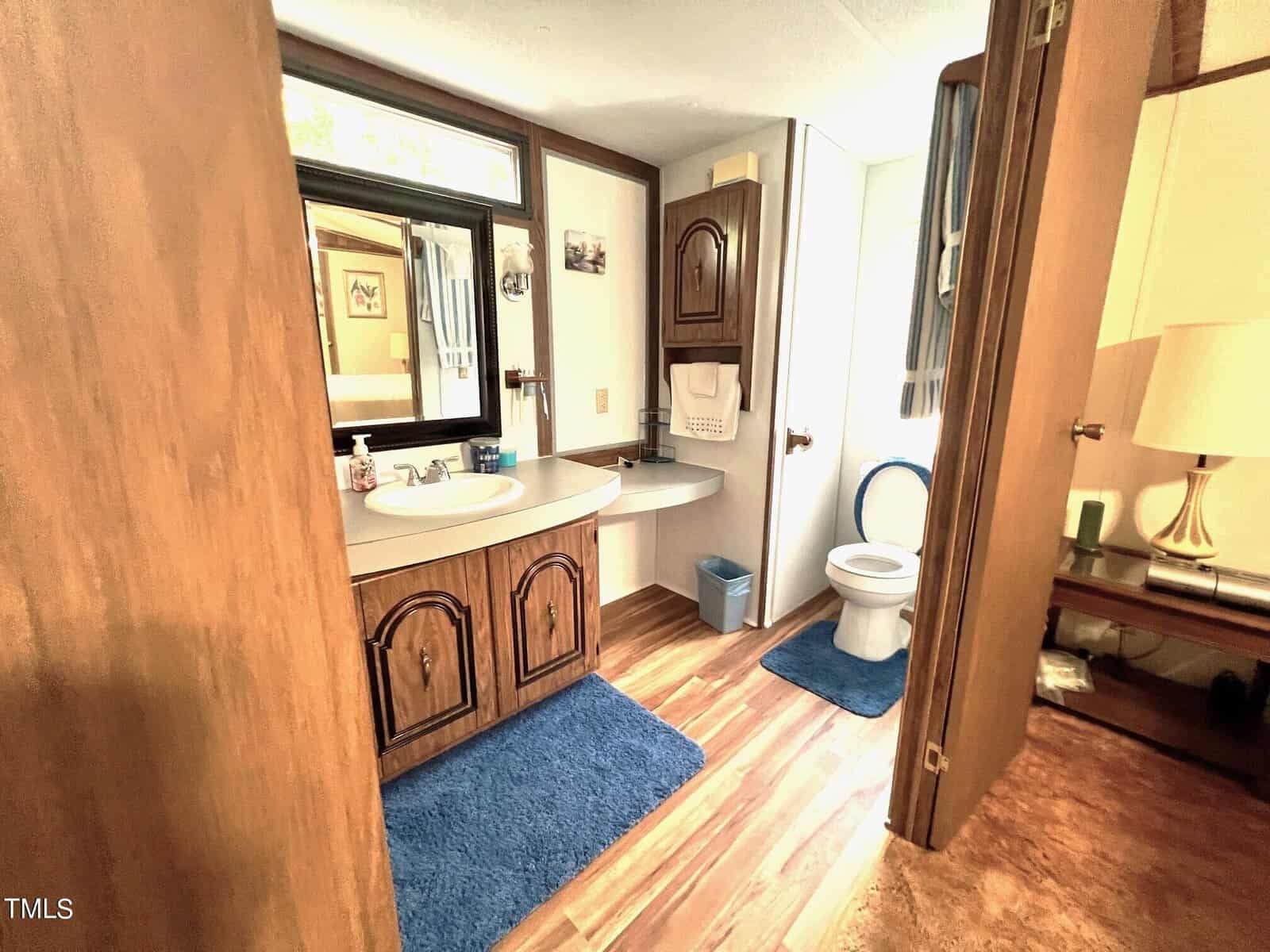 A bathroom with wooden cabinets, a white sink, and a toilet. Blue rugs are on the floor, and a small window is above the sink. The walls and flooring have a warm, wooden tone, creating a cozy atmosphere. A lamp is visible in the adjacent room.