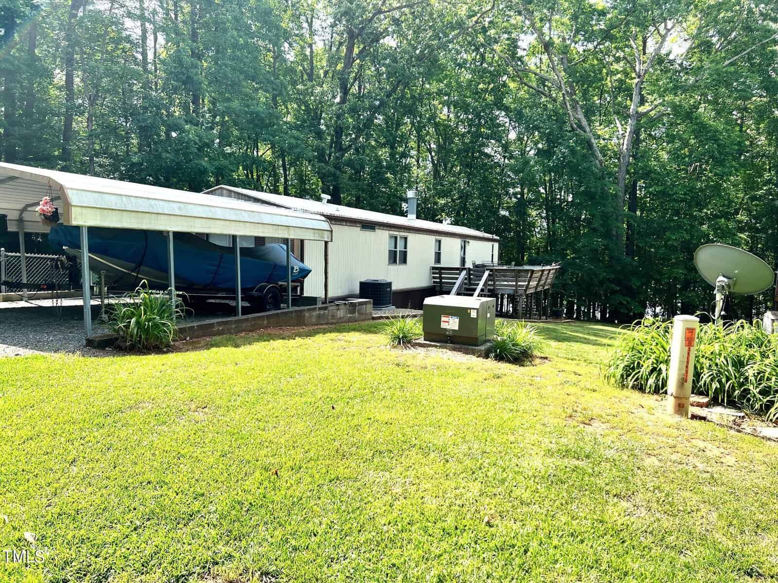 A white mobile home with a carport sheltering a boat is surrounded by trees. There's a satellite dish on the lawn, a metal structure with solar panels, and a green utility box nearby. The grass is well-maintained under a clear, sunny sky.