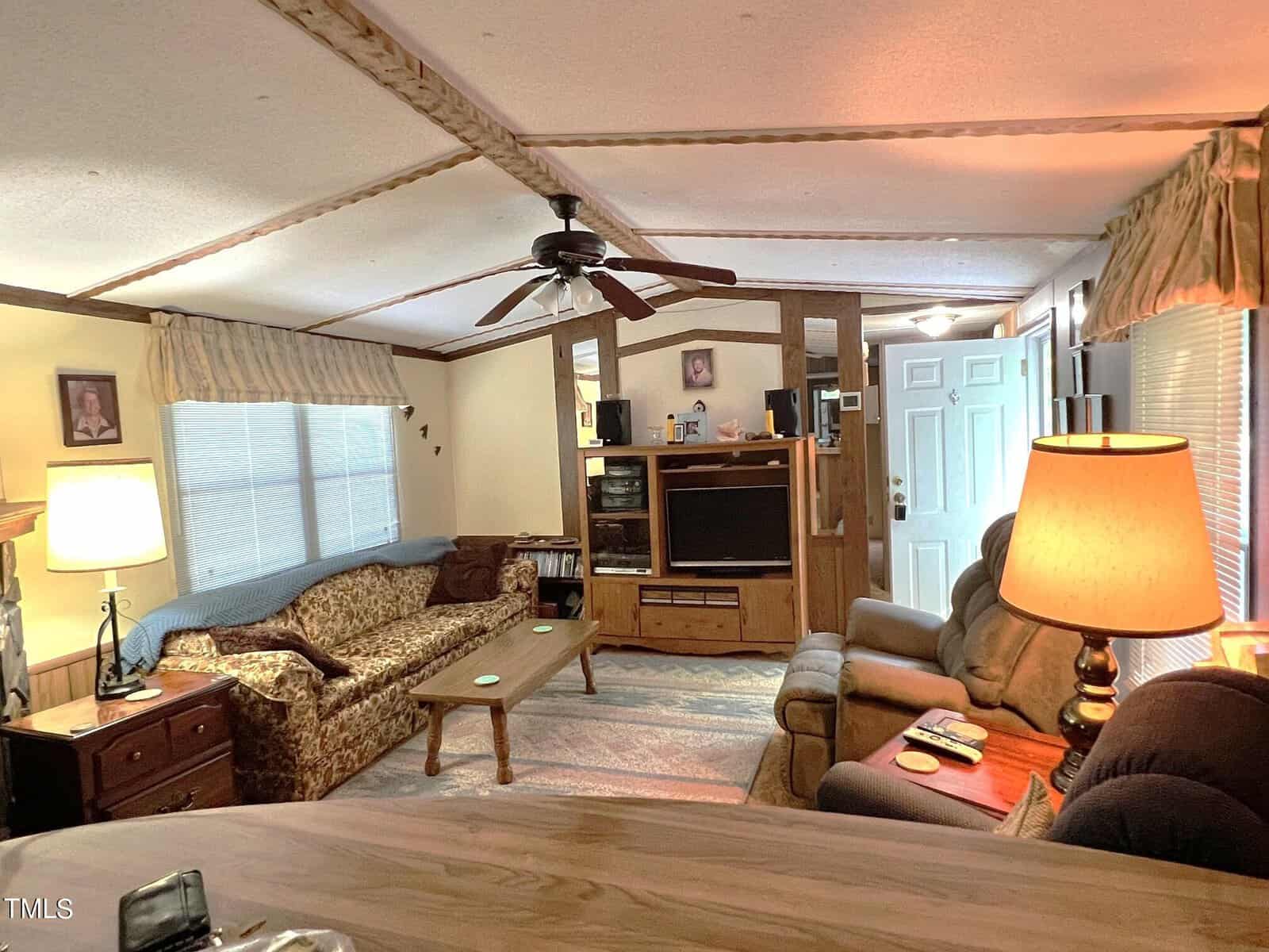 A cozy living room features a sofa with a patterned cover, a recliner, a wooden coffee table, a TV in an entertainment center, and a ceiling fan. The space is softly lit by a floor lamp and a table lamp near the entrance door.