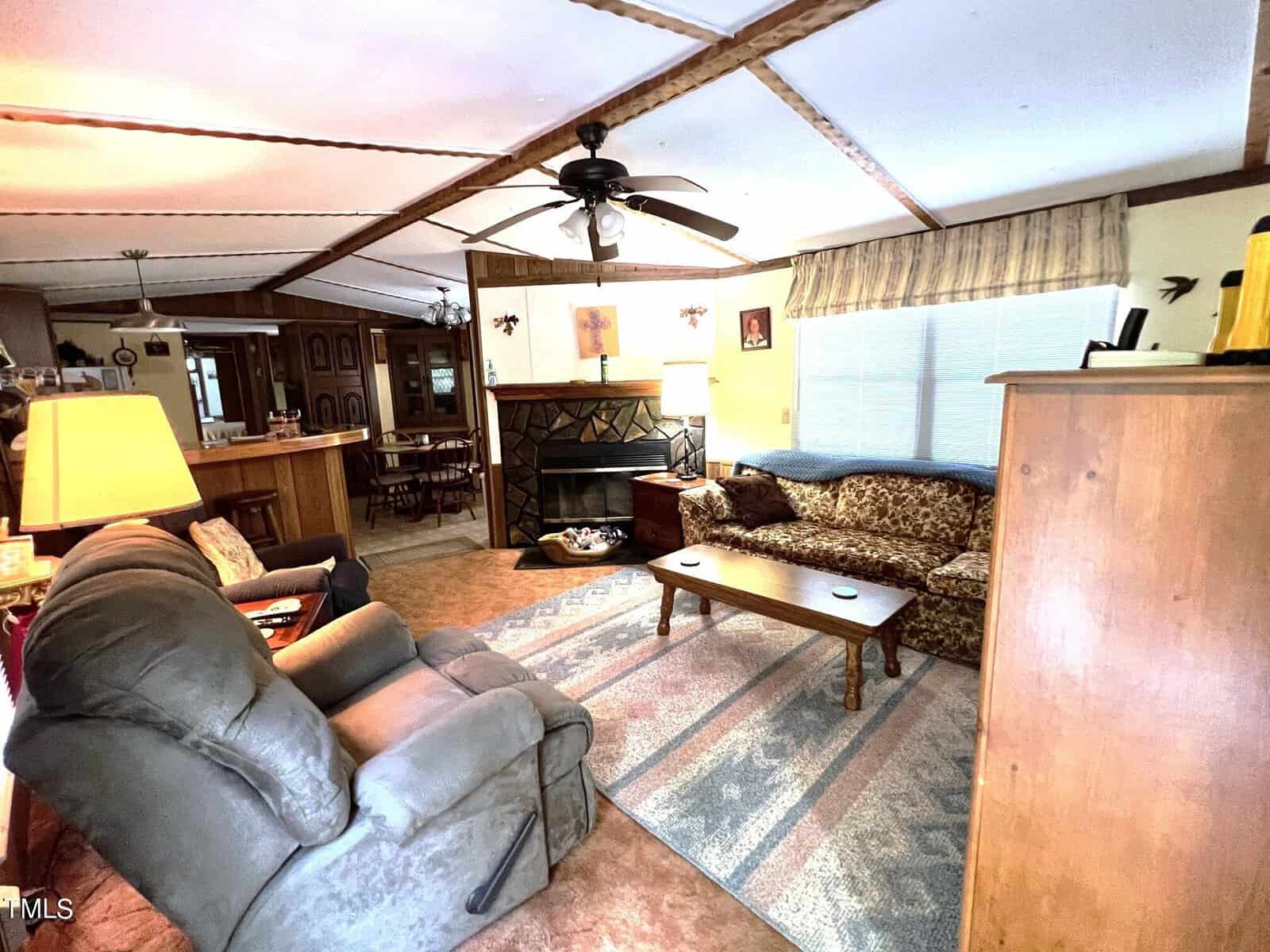 A cozy living room with a patterned sofa, two recliners, a wooden coffee table, and a fireplace. A ceiling fan hangs above, and there are a few paintings on the wall. Sunlight streams in through a window with striped curtains.