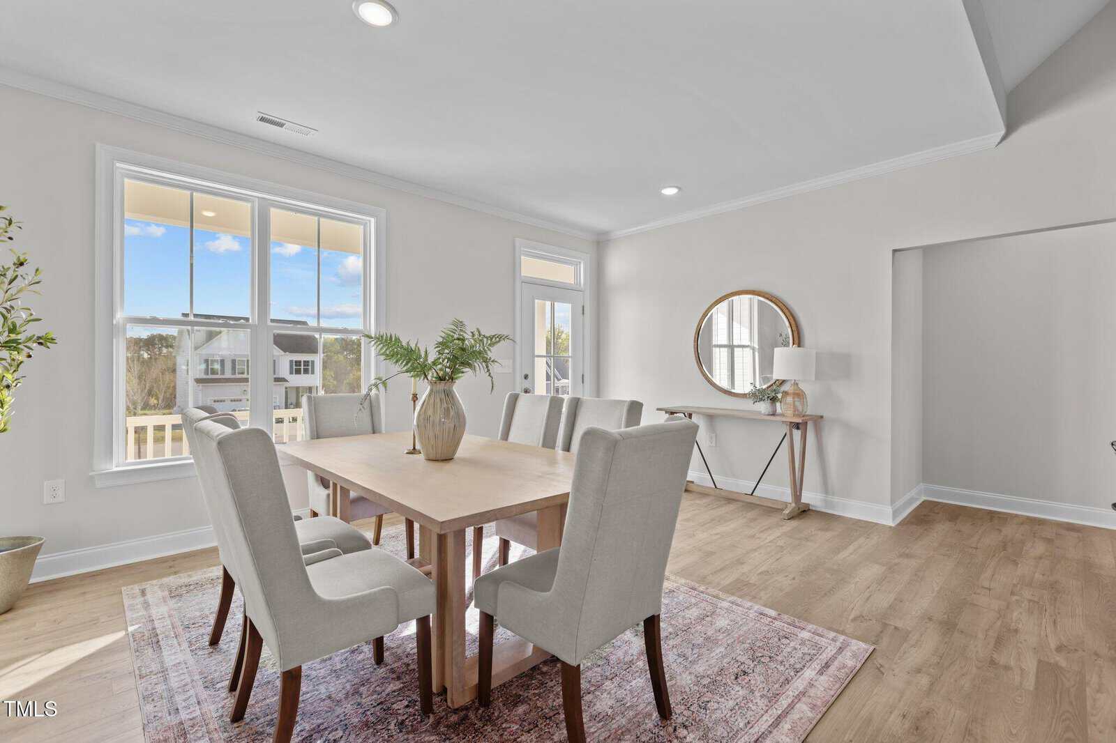 Bright dining room with a light wood table, six upholstered chairs, large window, round mirror above a console table, and hardwood floors. The space is decorated with green plants and has natural light streaming in.
