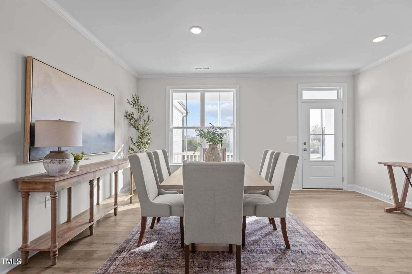 Bright dining room with a rectangular wooden table, six upholstered chairs, a large window, a glass door, a sideboard with a lamp and artwork, and a plant in the corner on a patterned rug.