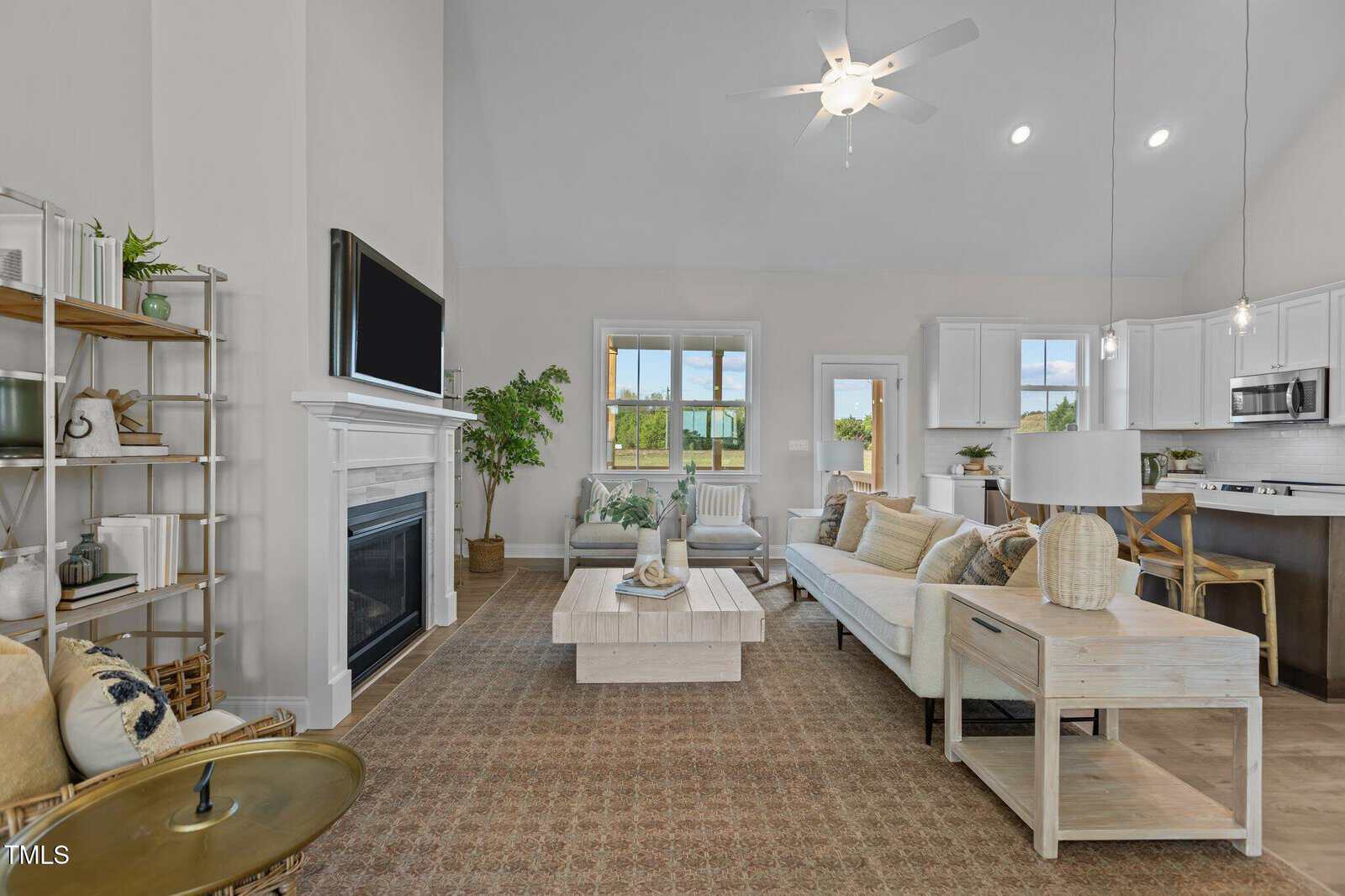 Bright living room with white walls, beige carpet, modern furniture, wall-mounted TV, fireplace, potted plant, open shelves, and large windows letting in natural light. Kitchen and dining area visible in the background.
