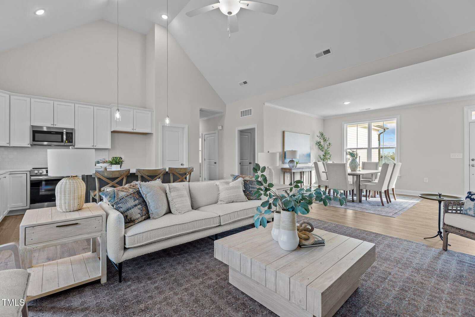 Modern open-concept living room and dining area with white walls, vaulted ceiling, neutral furnishings, kitchen in the background, and large windows letting in natural light.