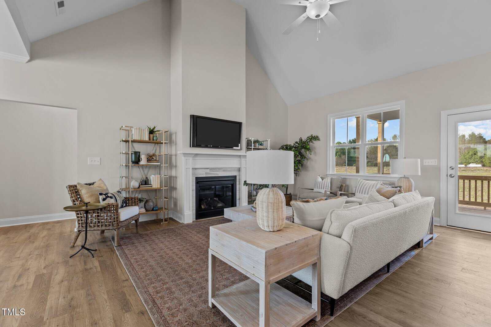 A modern living room with vaulted ceilings, light walls, wood floors, a white sectional sofa, a fireplace with a TV above, and large windows letting in natural light. There are accent chairs, shelves, and a glass door to the outside.