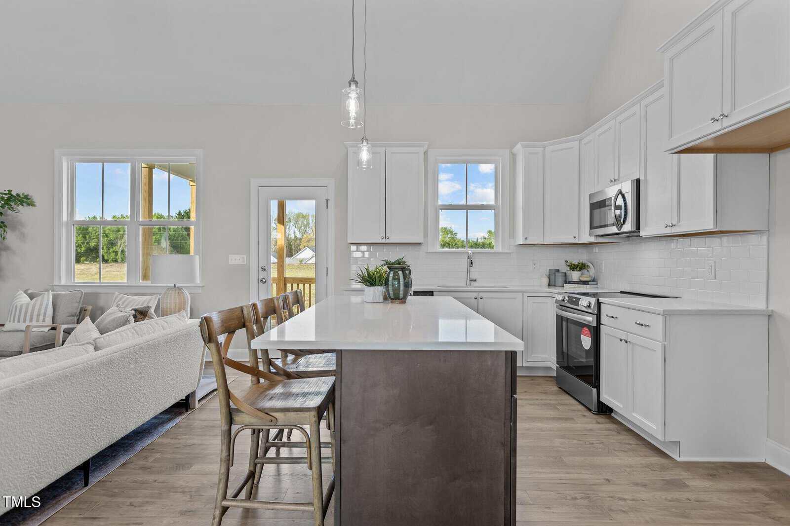 Modern kitchen with white cabinets, stainless steel appliances, and a large island with wooden chairs. Bright space with large windows, natural light, and an open view into a cozy living room.
