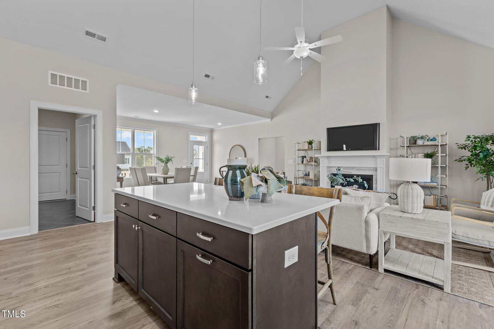 Modern open-concept living area with a kitchen island in the foreground, light wood flooring, neutral-toned furniture, a fireplace, wall-mounted TV, ceiling fan, and large windows letting in natural light.