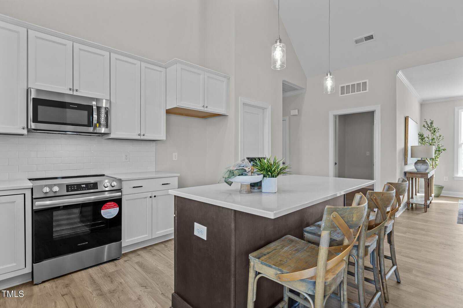 Modern kitchen with white cabinets, stainless steel appliances, and a large island with a sink. Three wooden bar stools line the island, and decorative plants are arranged on the countertop.