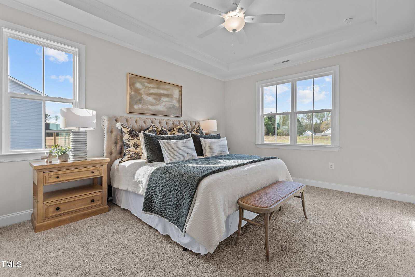 Bright bedroom with a large bed, neutral bedding, two nightstands with lamps, a wooden bench at the foot of the bed, beige carpet, and windows letting in natural light.