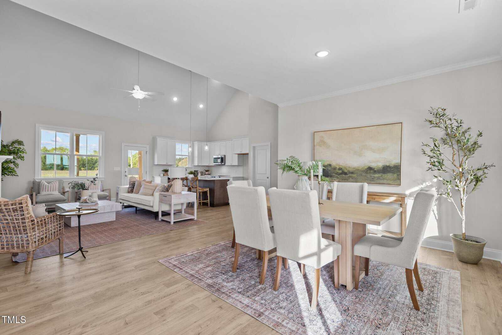 Bright, open-concept living and dining area with light-colored walls, wood flooring, neutral furniture, large windows, a dining table with six chairs, modern kitchen, and decorative plants.