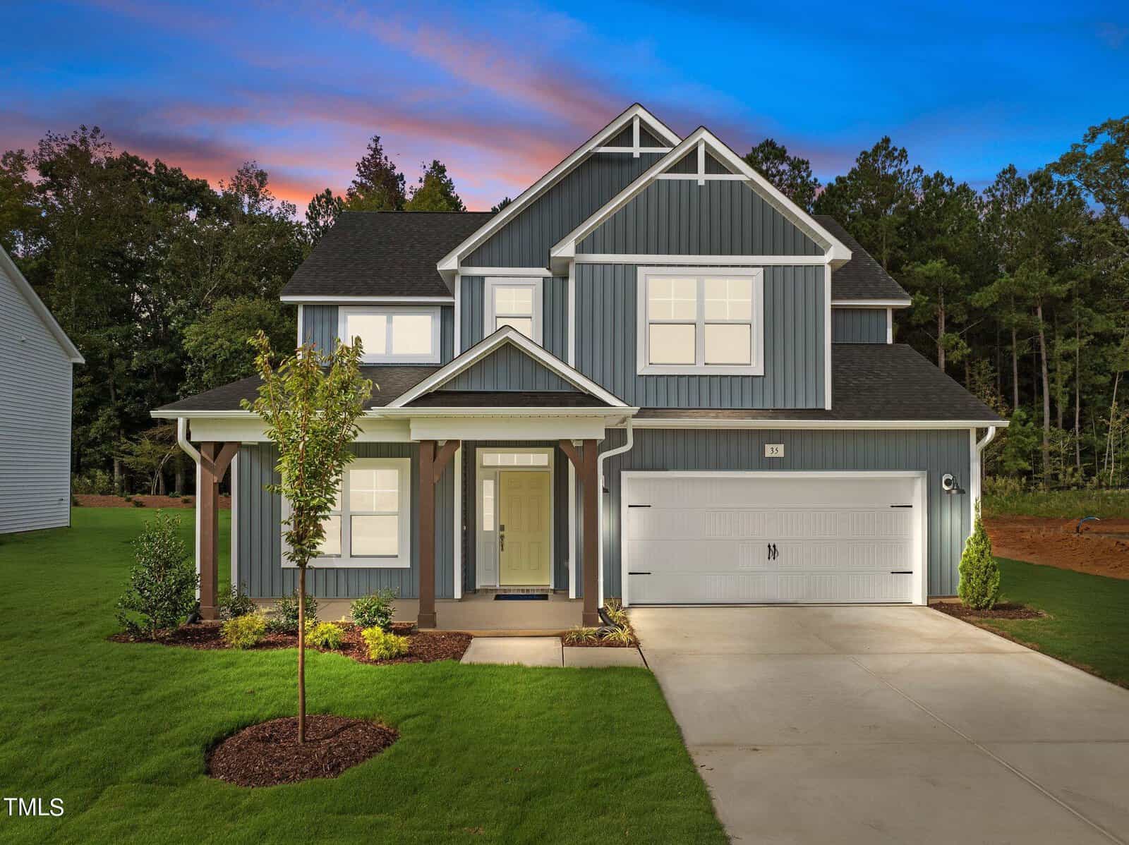 A modern two-story house with blue siding, white trim, a covered front porch, double garage, and landscaped lawn, set against a twilight sky and surrounded by trees.
