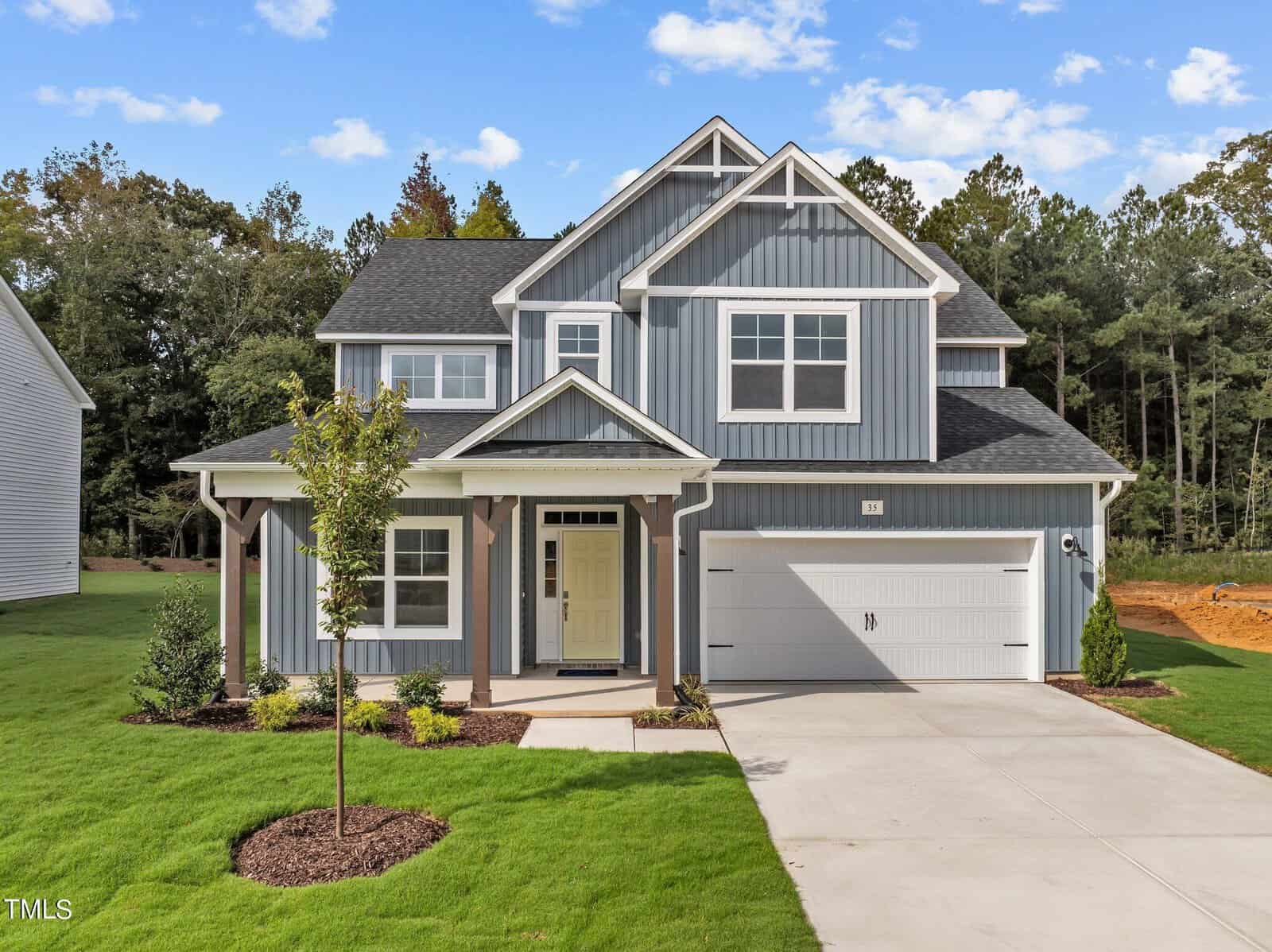 A modern two-story blue house with white trim, a front porch, attached two-car garage, manicured lawn, and young trees, set against a backdrop of trees under a blue sky with scattered clouds.