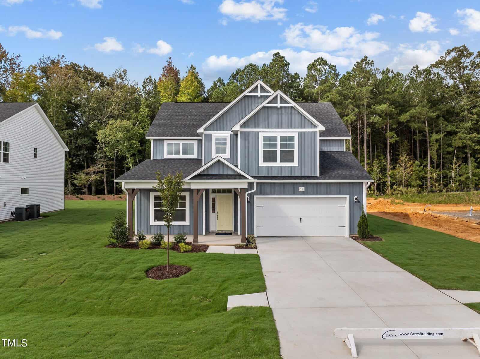 A two-story modern gray house with white trim, a double garage, and a well-manicured lawn sits on a spacious lot with trees and new construction in the background under a partly cloudy sky.