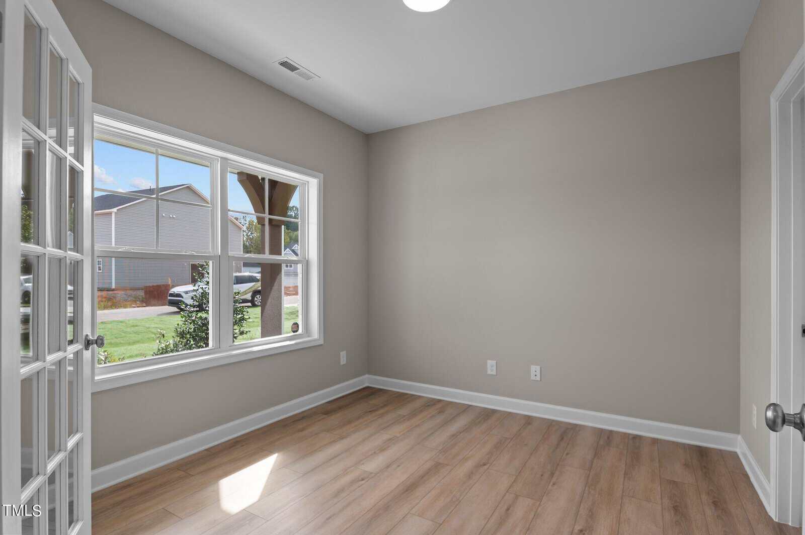 A small, empty room with light wood flooring, beige walls, a large window letting in natural light, and glass French doors. The view through the window shows a suburban street with houses and greenery.