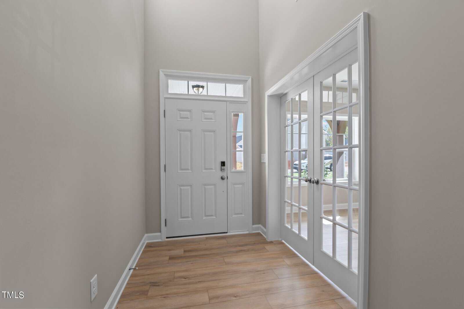 A bright entryway with light-colored walls and wood flooring features a white front door with glass panels. To the right, double French doors open to a sunlit room with large windows.