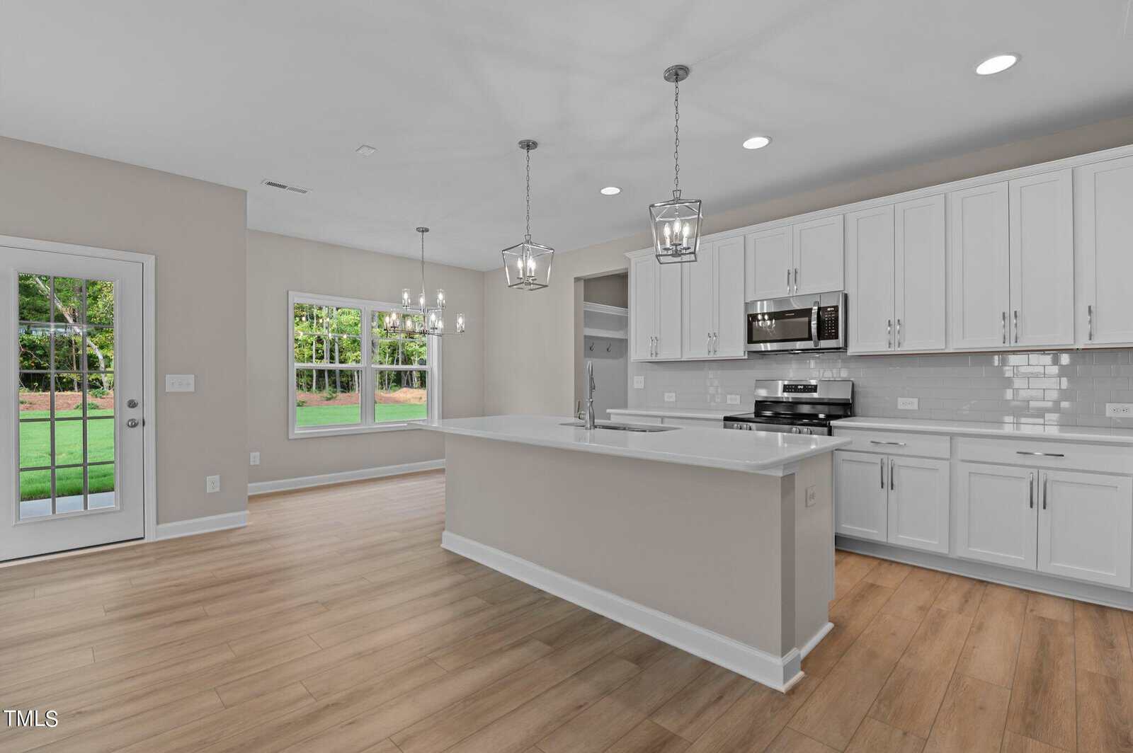 A modern kitchen with white cabinets, stainless steel appliances, a large island with pendant lights, and light wood flooring. A dining area with a chandelier and large windows overlooks a green backyard.